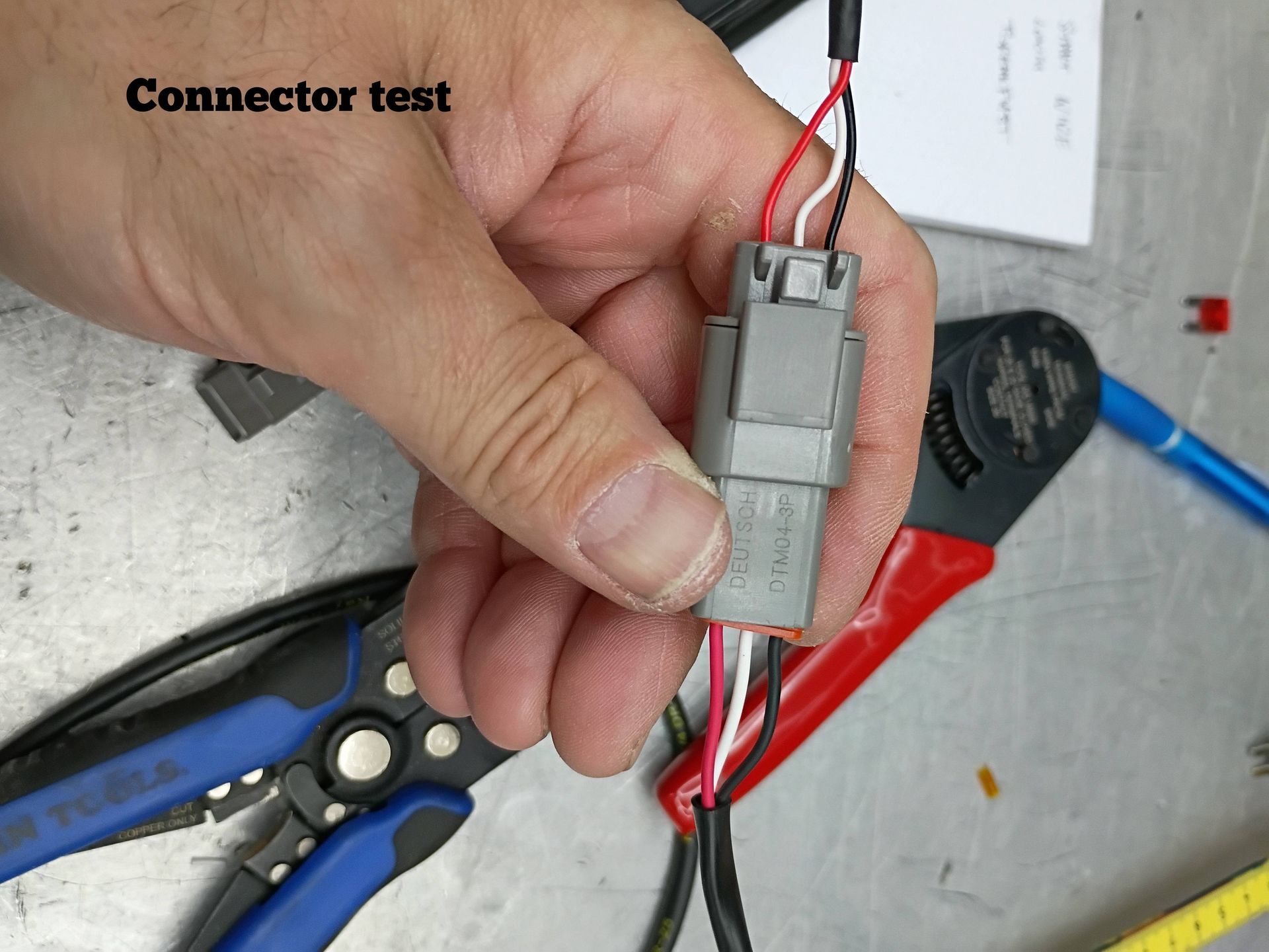Hand holding gray electrical connector with red and black wires on a workbench, with tools nearby