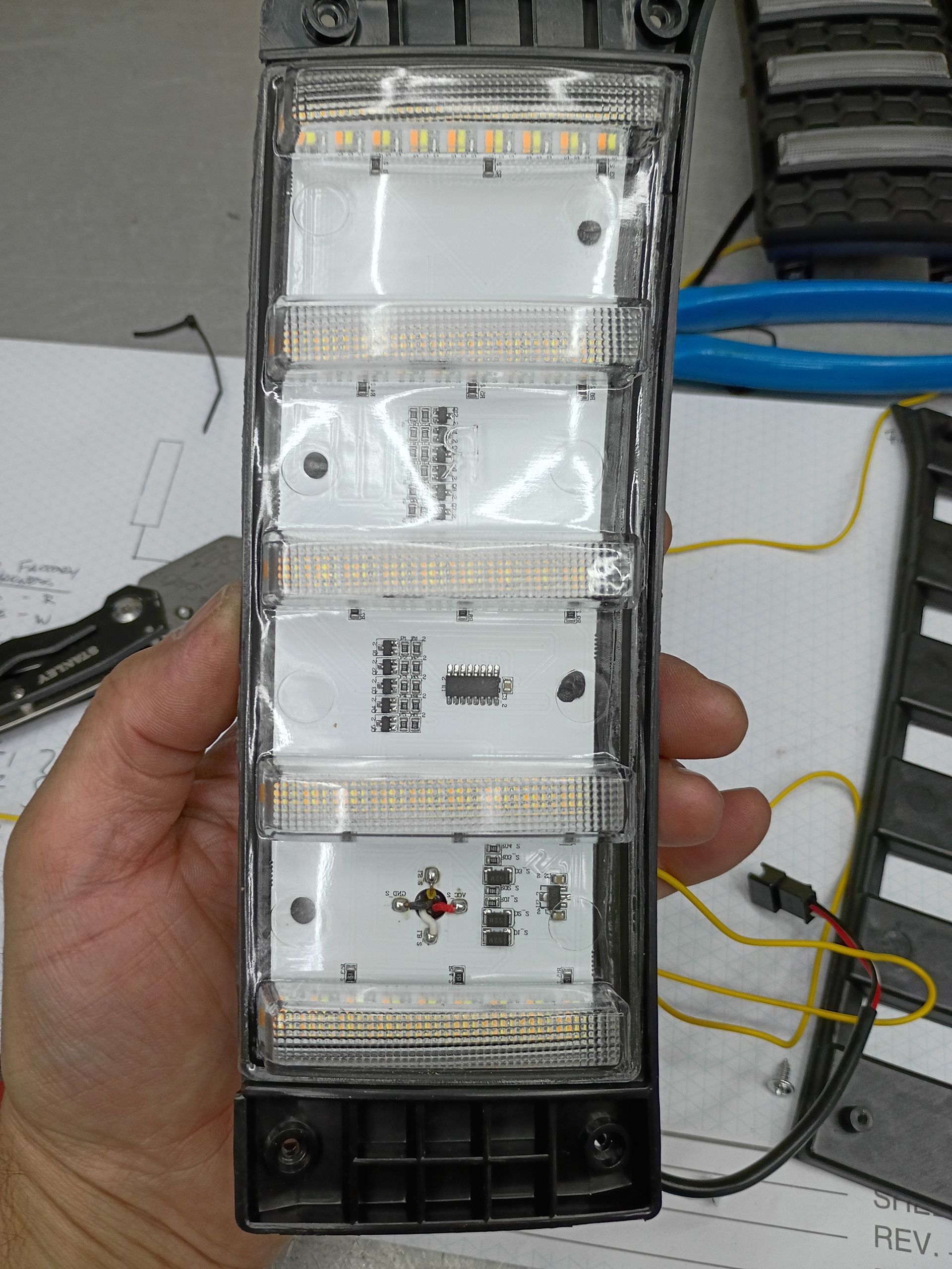 Hand holding a tall clear plastic device with stacked modules and wires on a workbench