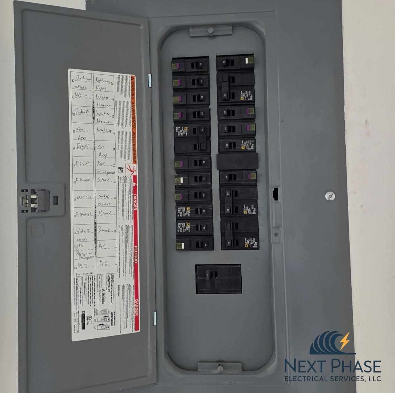 Gray electrical panel with breakers, open door, and labeling on wall.