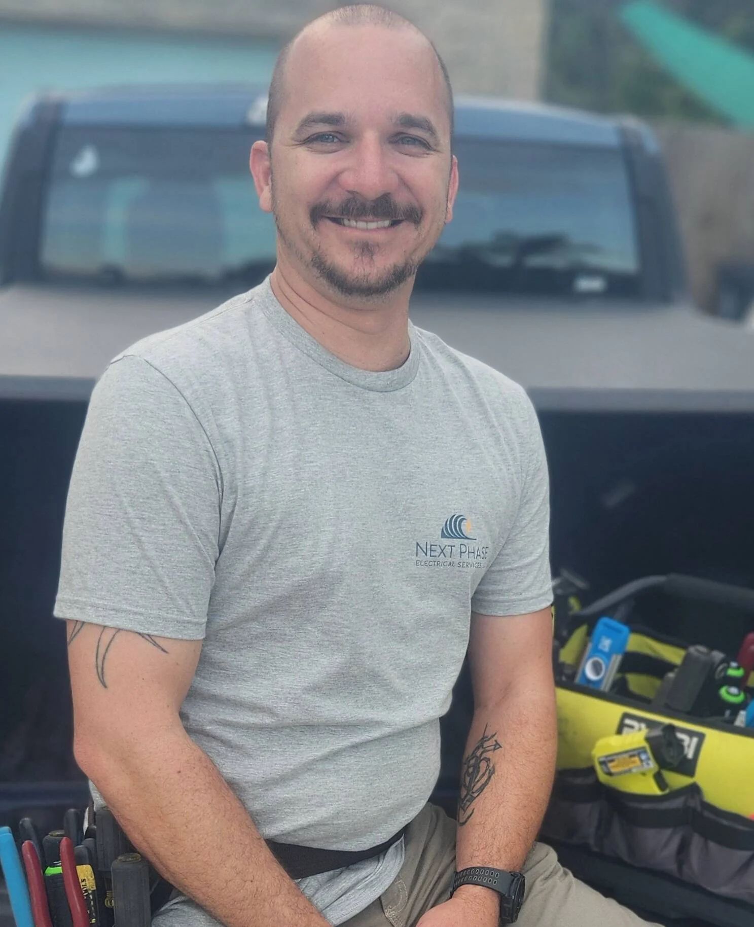 Man with mustache, gray t-shirt, smiling, seated in truck bed with tools.