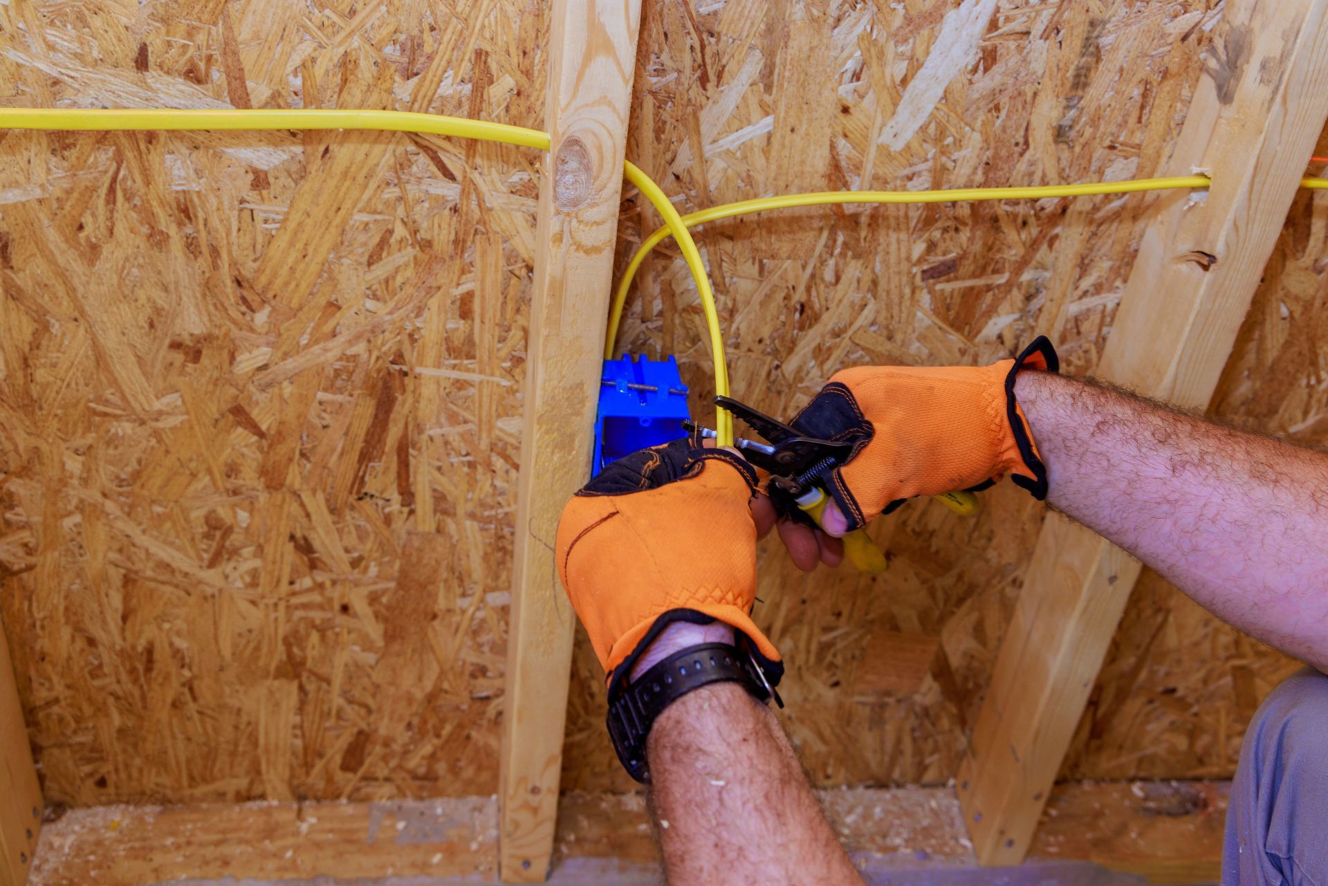 Person in orange gloves wiring yellow electrical cables in an unfinished wooden attic.