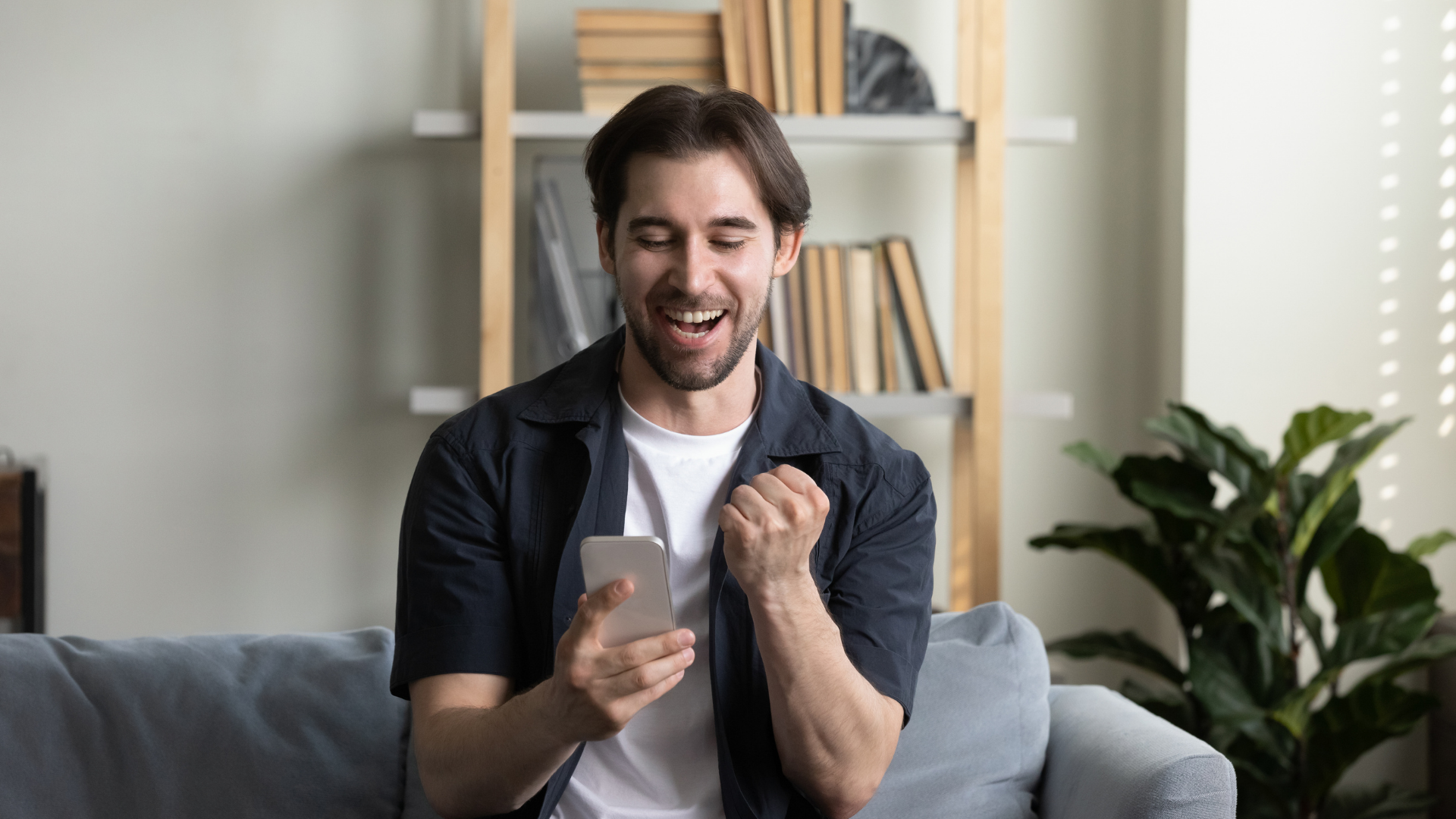 Man smiling on a sofa, holding a phone and celebrating, in a bright living room