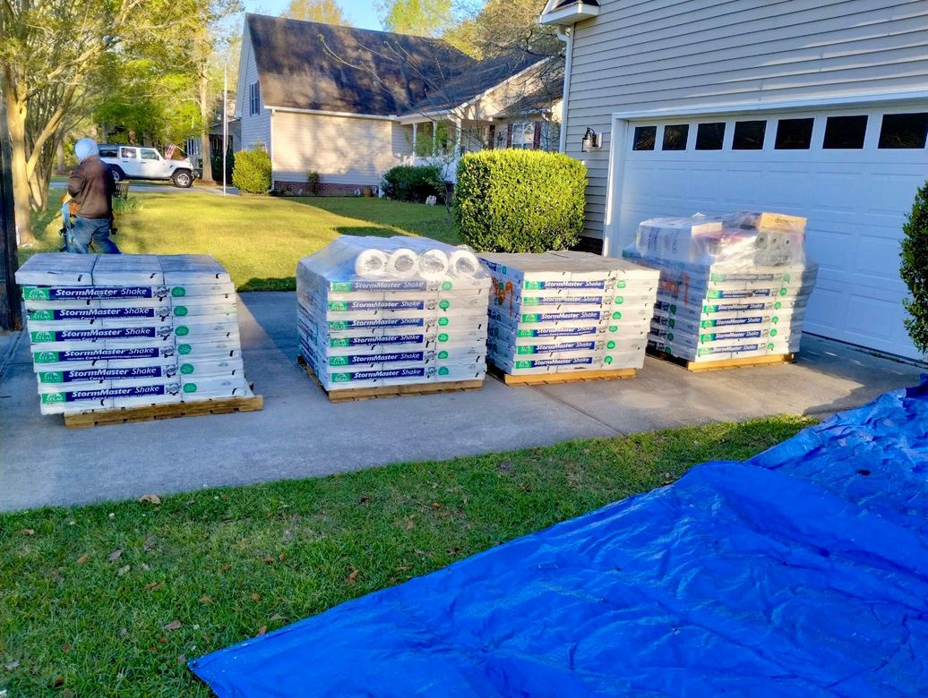 A man is standing in front of a house next to a blue tarp with pallets of roof singles