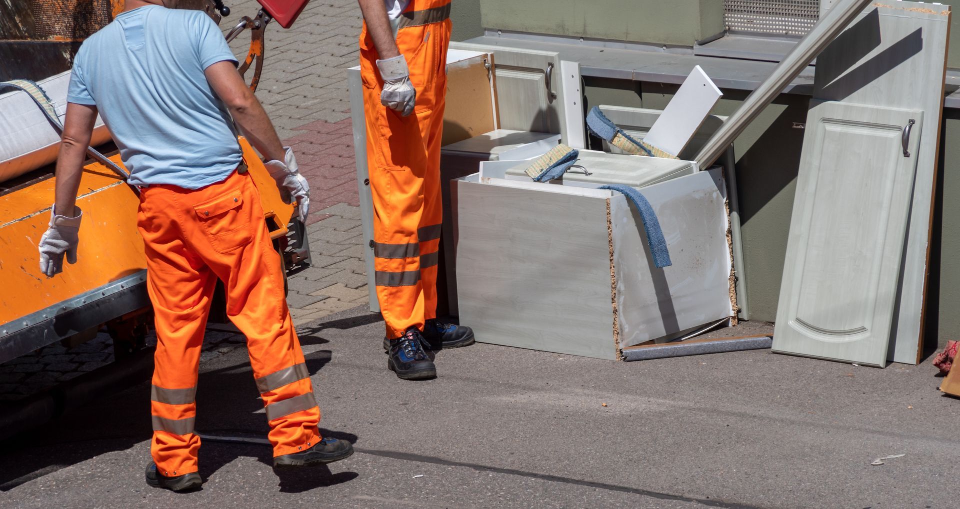 Two sanitation workers in orange uniforms loading discarded cabinet parts onto a truck.