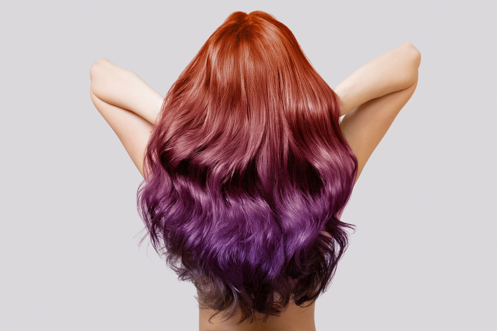 Back view of long wavy hair dyed red fading to purple, with arms raised behind the head