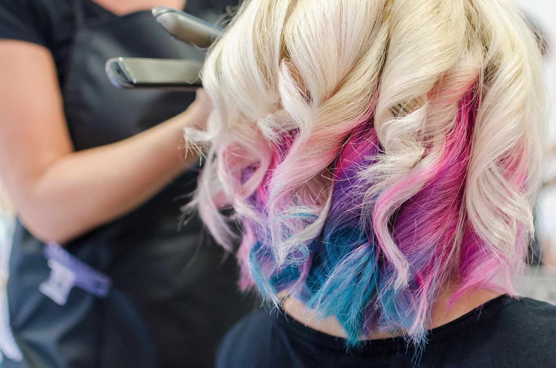 Blonde hair styled in curls with pink and teal streaks while being styled in a salon
