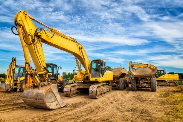 Yellow excavators and dump trucks parked on a dirt construction site under a cloudy blue sky.