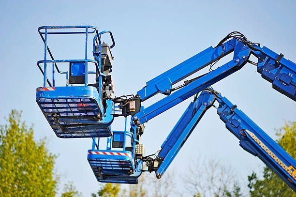 Two blue aerial work platforms, one elevated above the other, against a clear blue sky and trees.