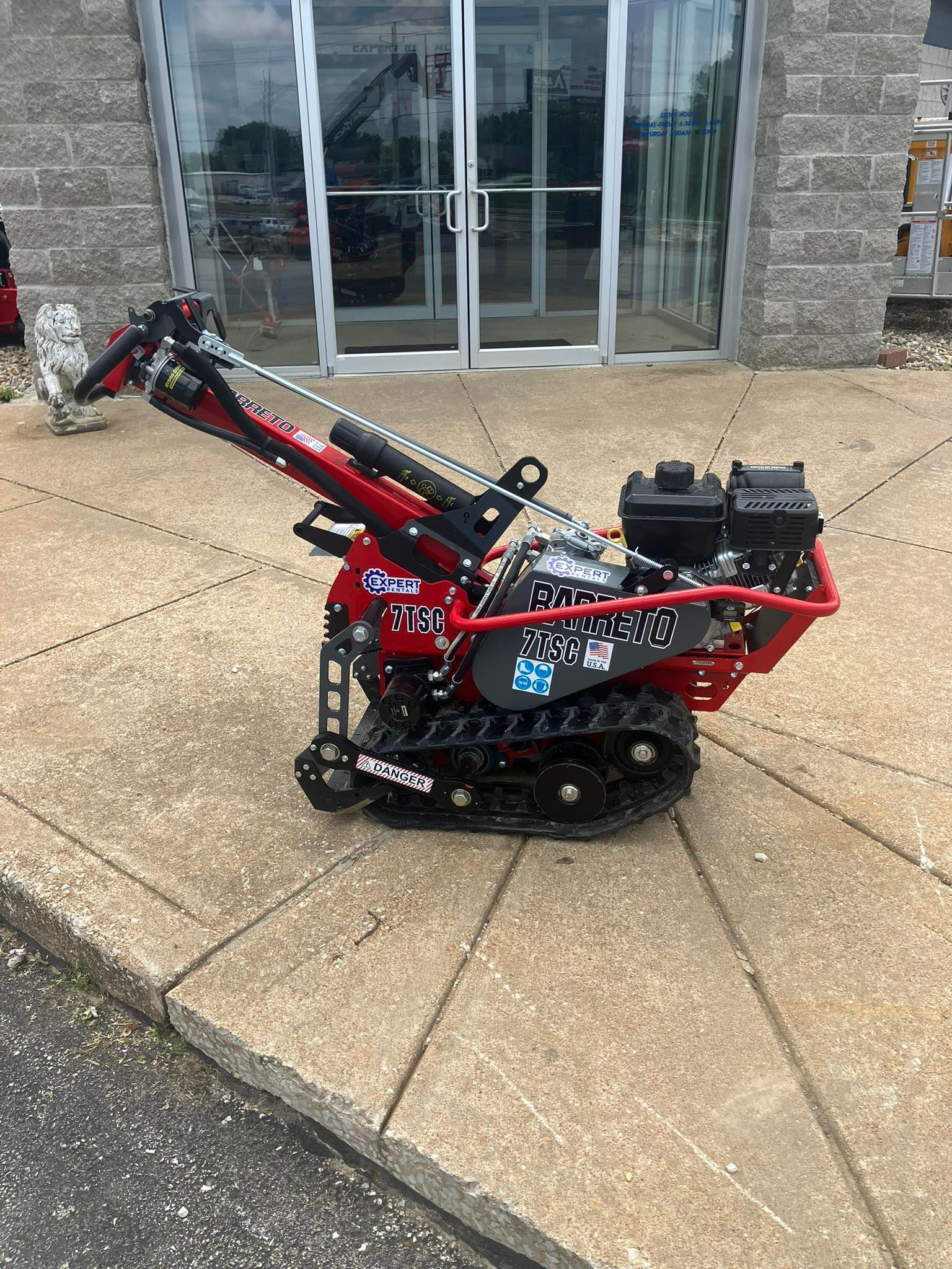 A red track-driven stump grinder sitting on a paved surface in front of a glass-door building.