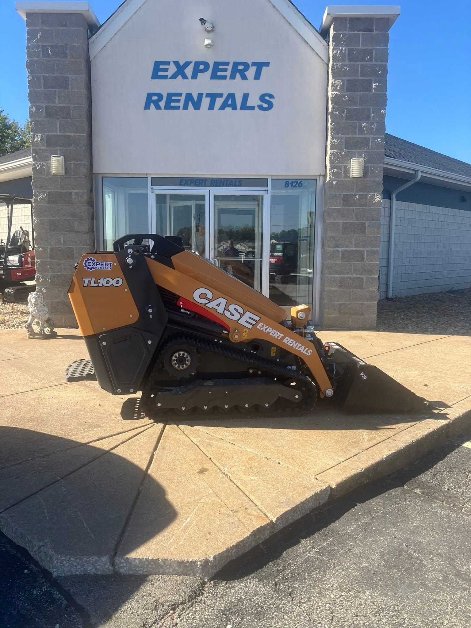 A tan and black Case brand skid steer loader parked on a sidewalk in front of an Expert Rentals store.
