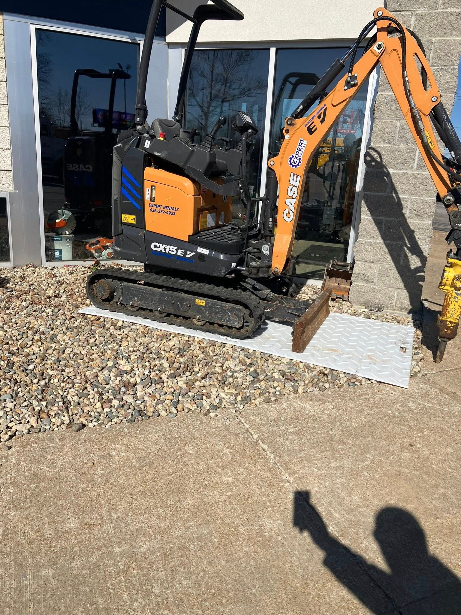 A Case brand mini-excavator with an orange arm and black body parked on a white mat on gravel outside a building.