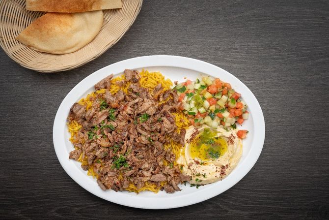Beef Shawarma Plate A white plate topped with meat , rice , hummus and vegetables.