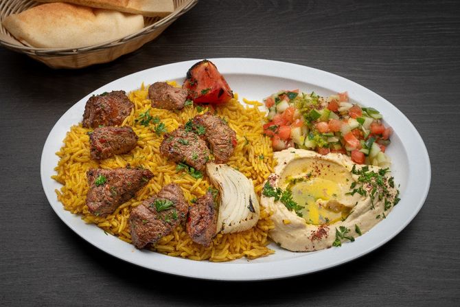 Beef Shish Kabab Plate A plate of food with rice , meat , hummus and vegetables on a table.