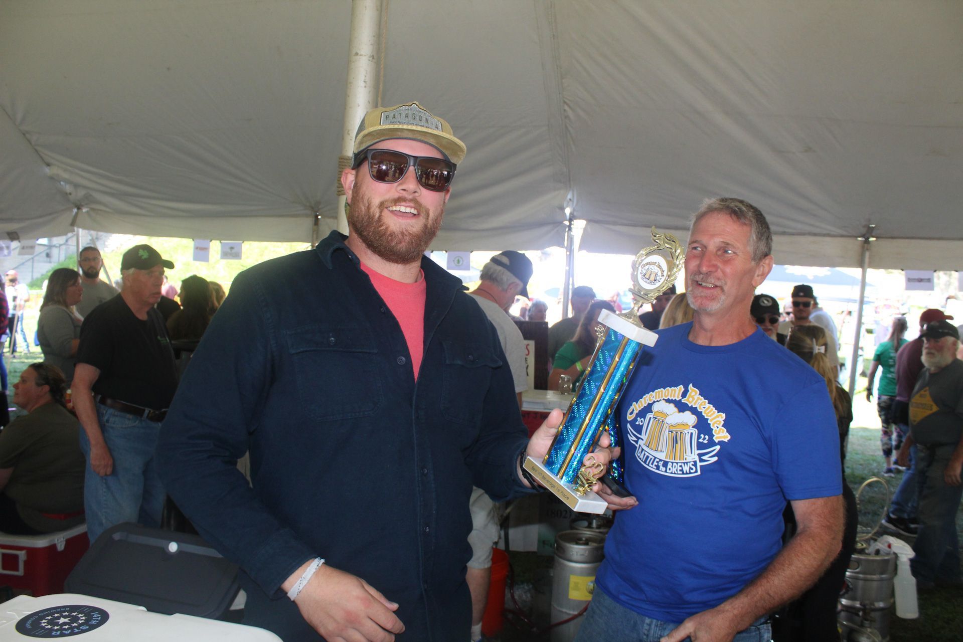 Two men are standing under a tent holding a trophy.