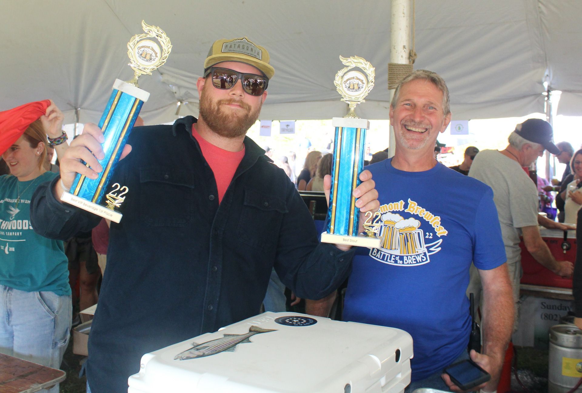 Two men are holding trophies in front of a white cooler.