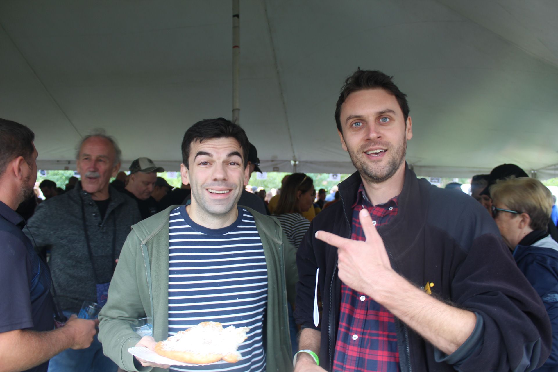 Two men are standing next to each other under a tent . one of the men is holding a plate of food.