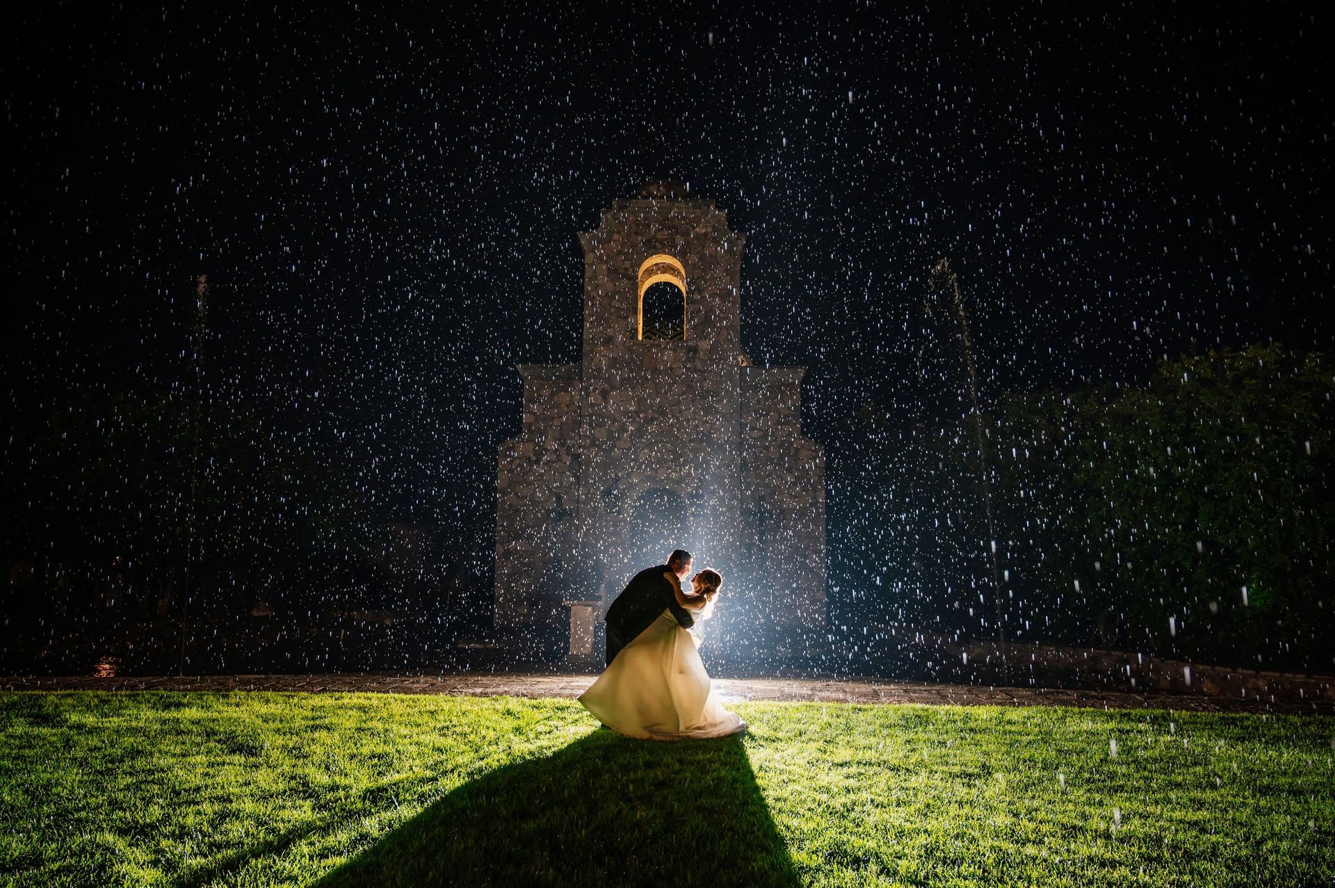 Novia de encaje y novio de traje negro comparten un abrazo íntimo en una habitación tenue.