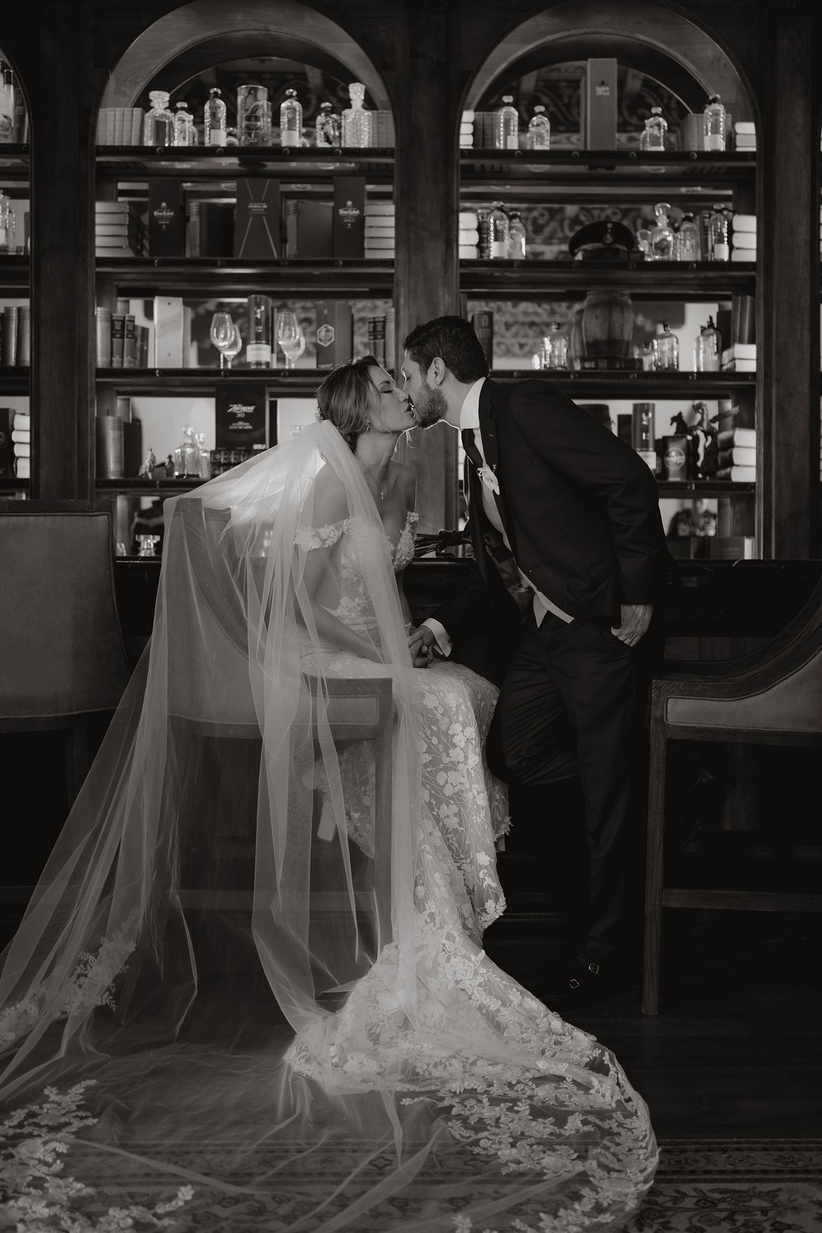 Una novia con un vestido de encaje y un largo velo, y un novio con traje, se besan frente a un bar con poca luz y estantes llenos de botellas.