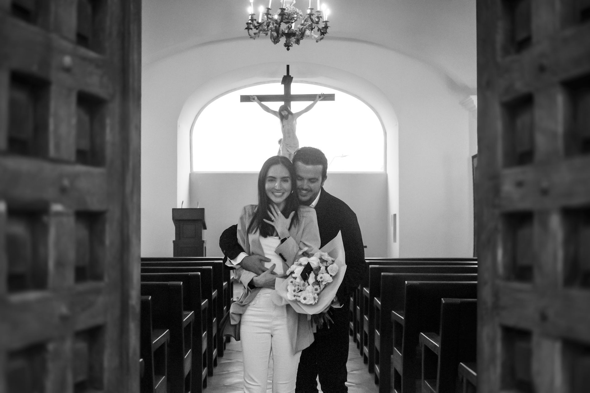 Una pareja se abraza en el pasillo de una iglesia, vista a través de una puerta, con un crucifijo visible al fondo.