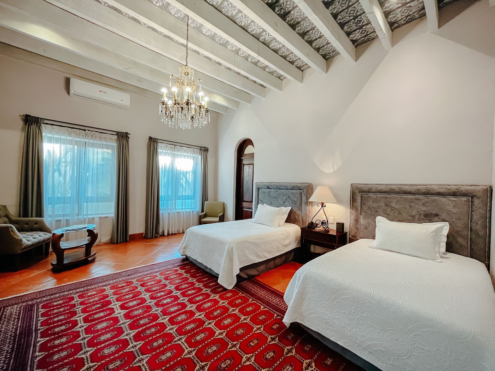 Una habitación de hotel con dos camas, una gran alfombra roja estampada, paredes blancas, vigas de techo a la vista y una lámpara de araña de cristal.
