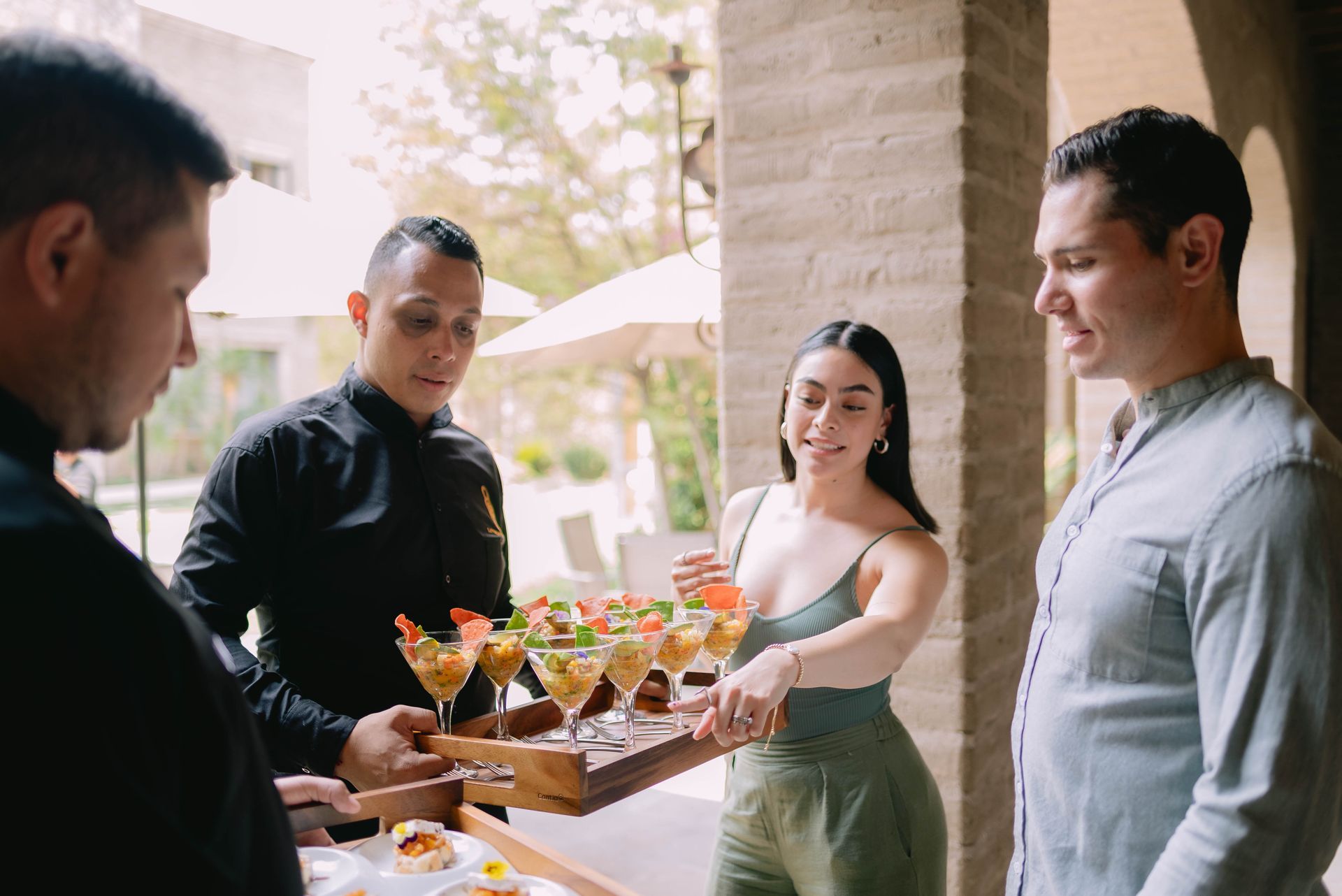 Una persona con una camisa negra sostiene una bandeja con pequeños cócteles mientras una mujer elige uno, de pie junto a otra persona.