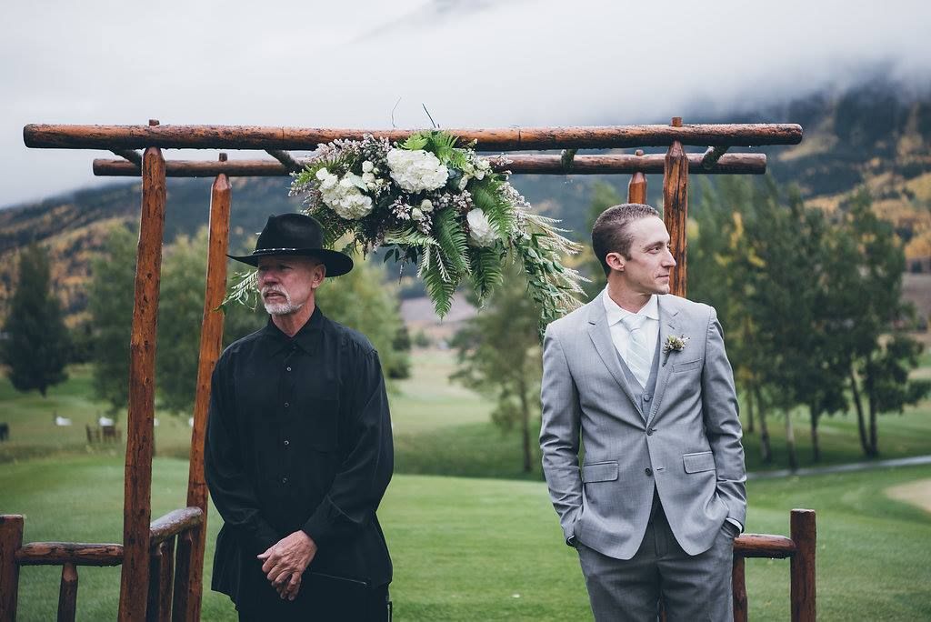 A groom and his father are standing under a wooden arch.