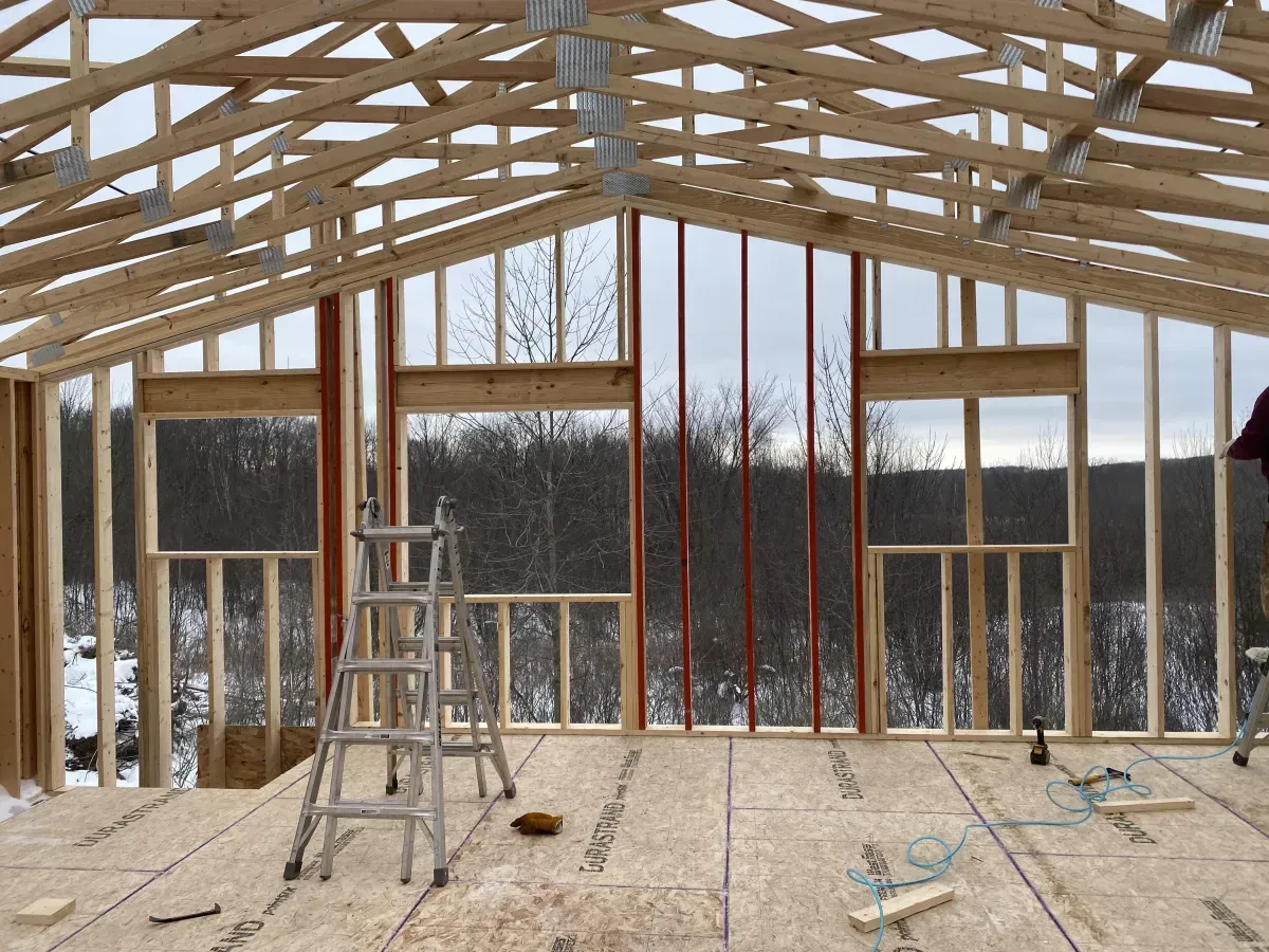 Interior of a wooden frame house under construction with a ladder, exposed beams, and large window openings.