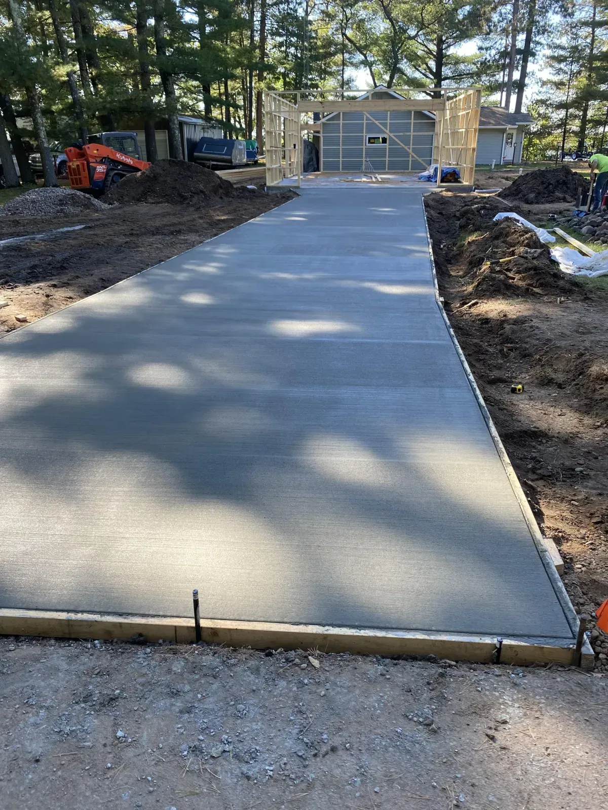 Concrete walkway under construction leading to a shed, with dirt piles and trees on both sides.