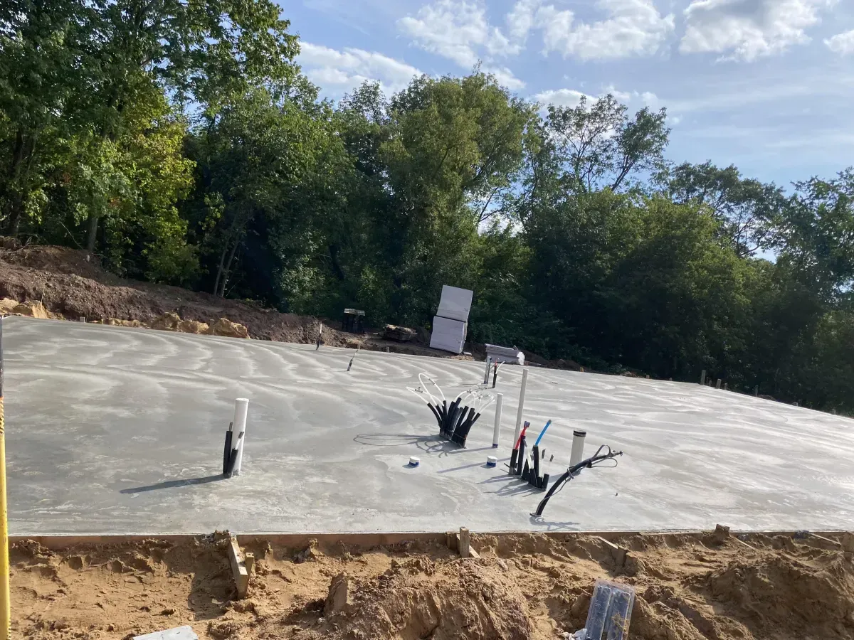 Concrete slab foundation under construction with exposed plumbing pipes and trees in the background