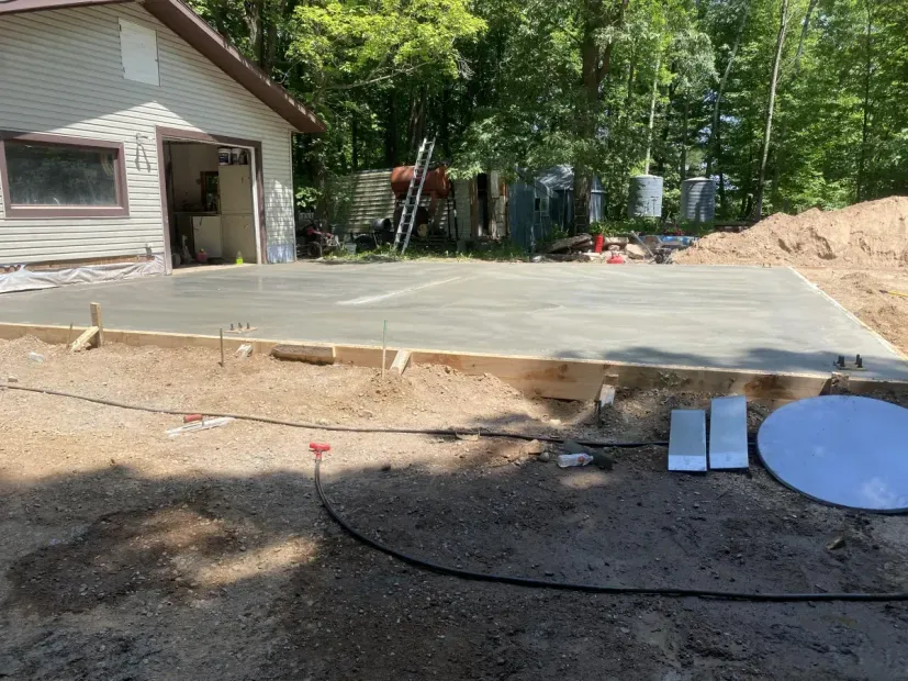 Fresh concrete driveway beside a house, with a dirt yard, trees, and construction materials nearby