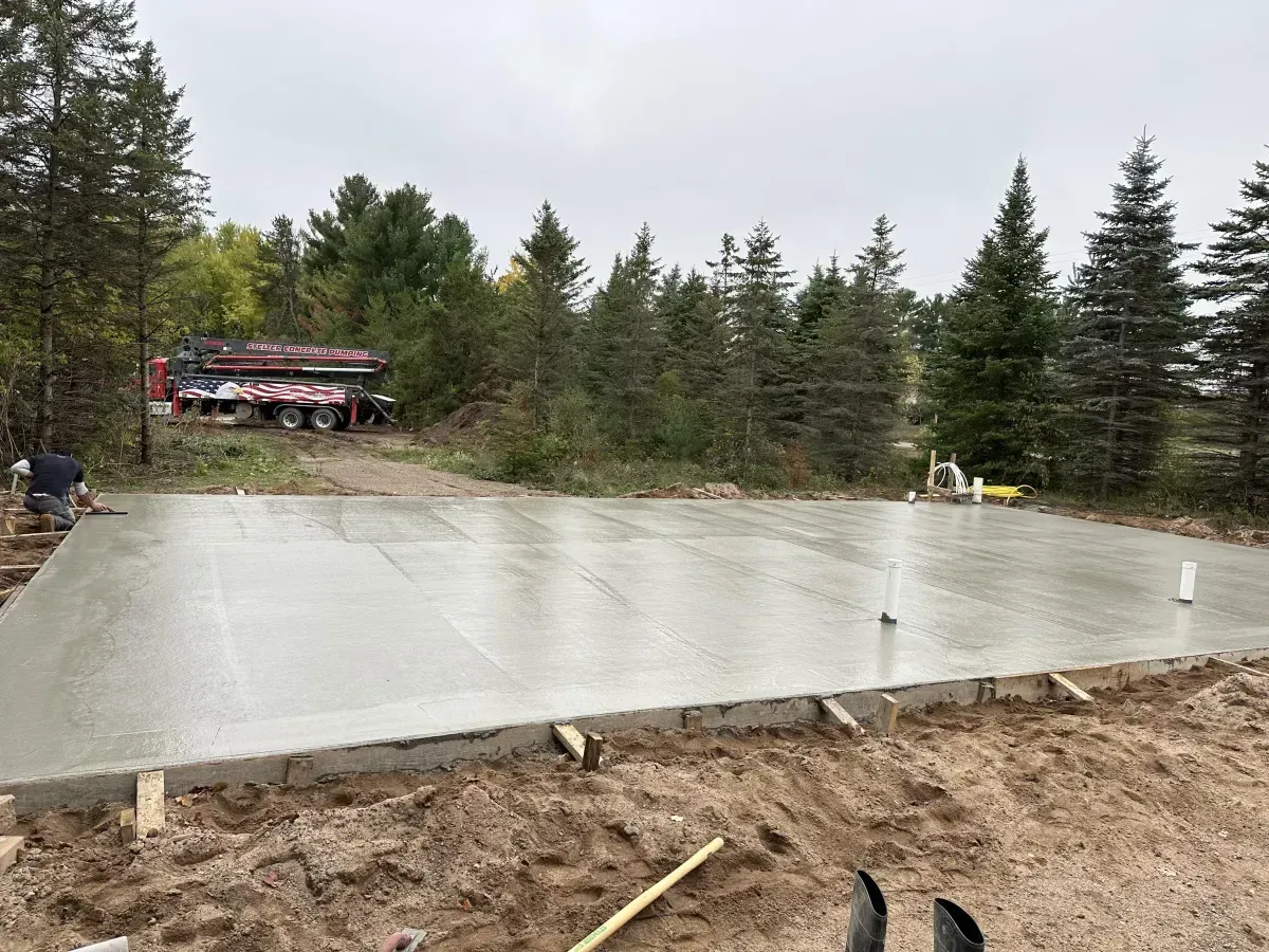 Freshly poured concrete foundation at a wooded construction site, with workers and a red building in the distance