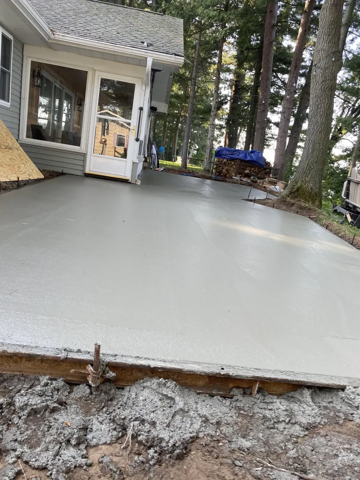 Freshly poured concrete patio beside a house and trees, with a doorway and blue tarp in the background
