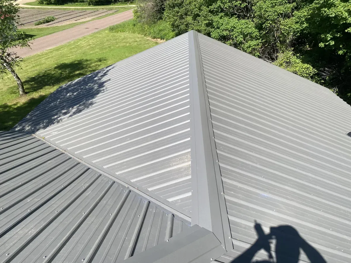 Gray metal roof viewed from above, with a sunlit yard and trees in the background.