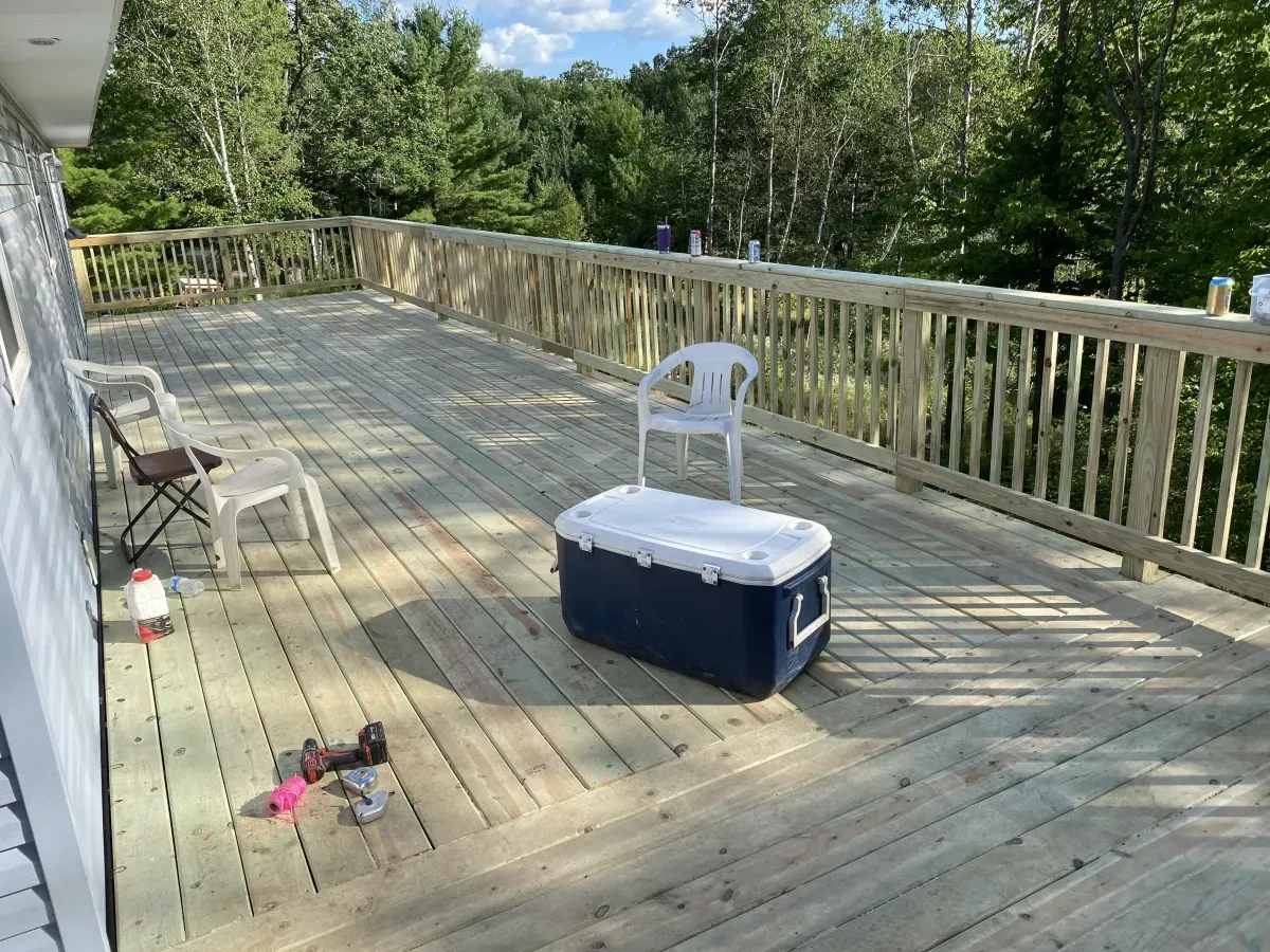Wooden deck with railings, cooler, chairs, and a few small items in sunlight.