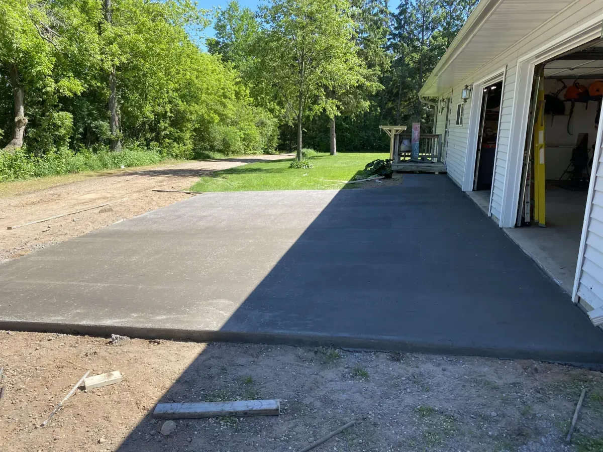 Concrete driveway slab beside a white garage, with trees and a dirt yard in the background.
