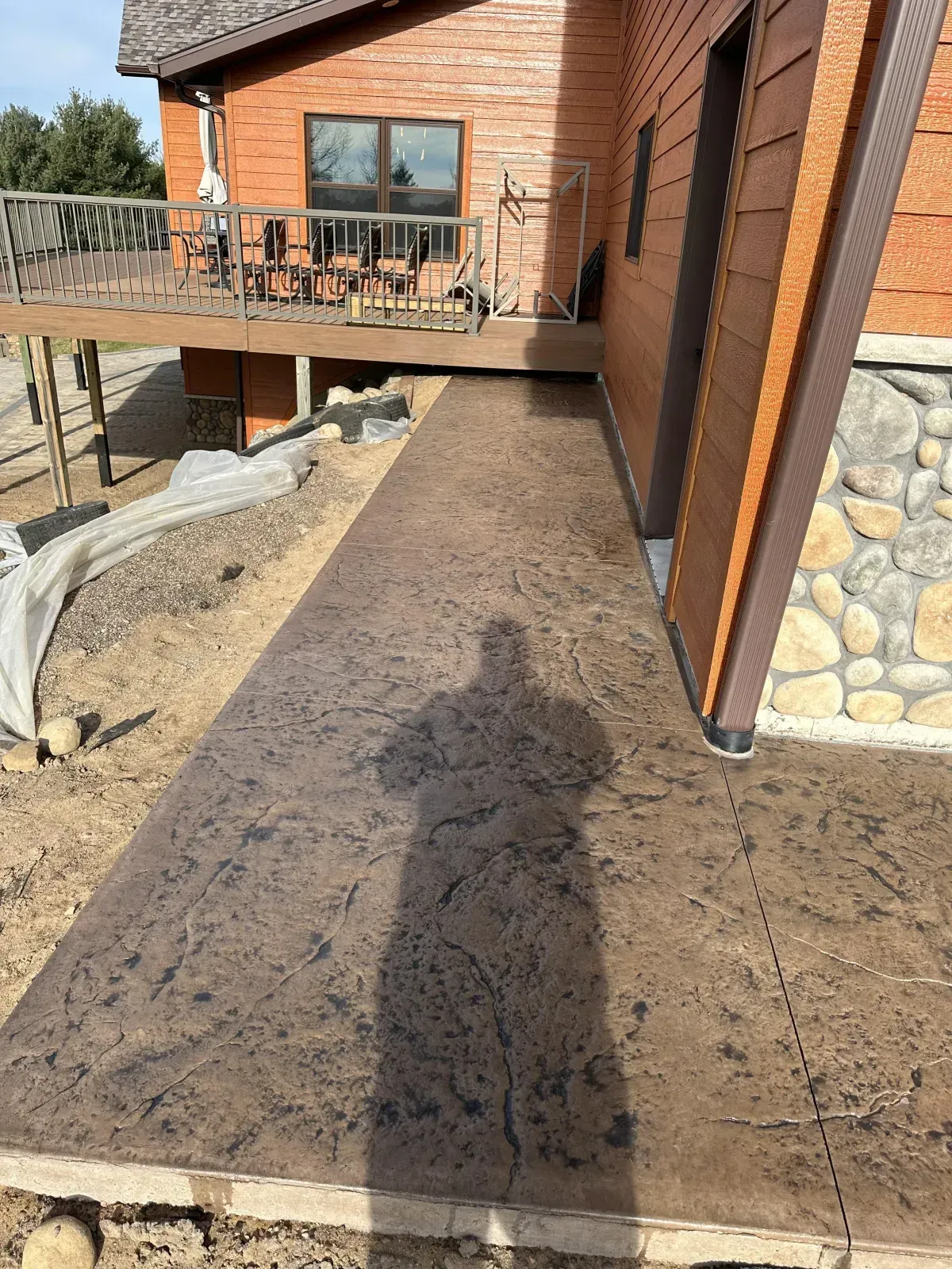 Concrete walkway beside a brick house, leading to a deck and window, with stone landscaping along the side.