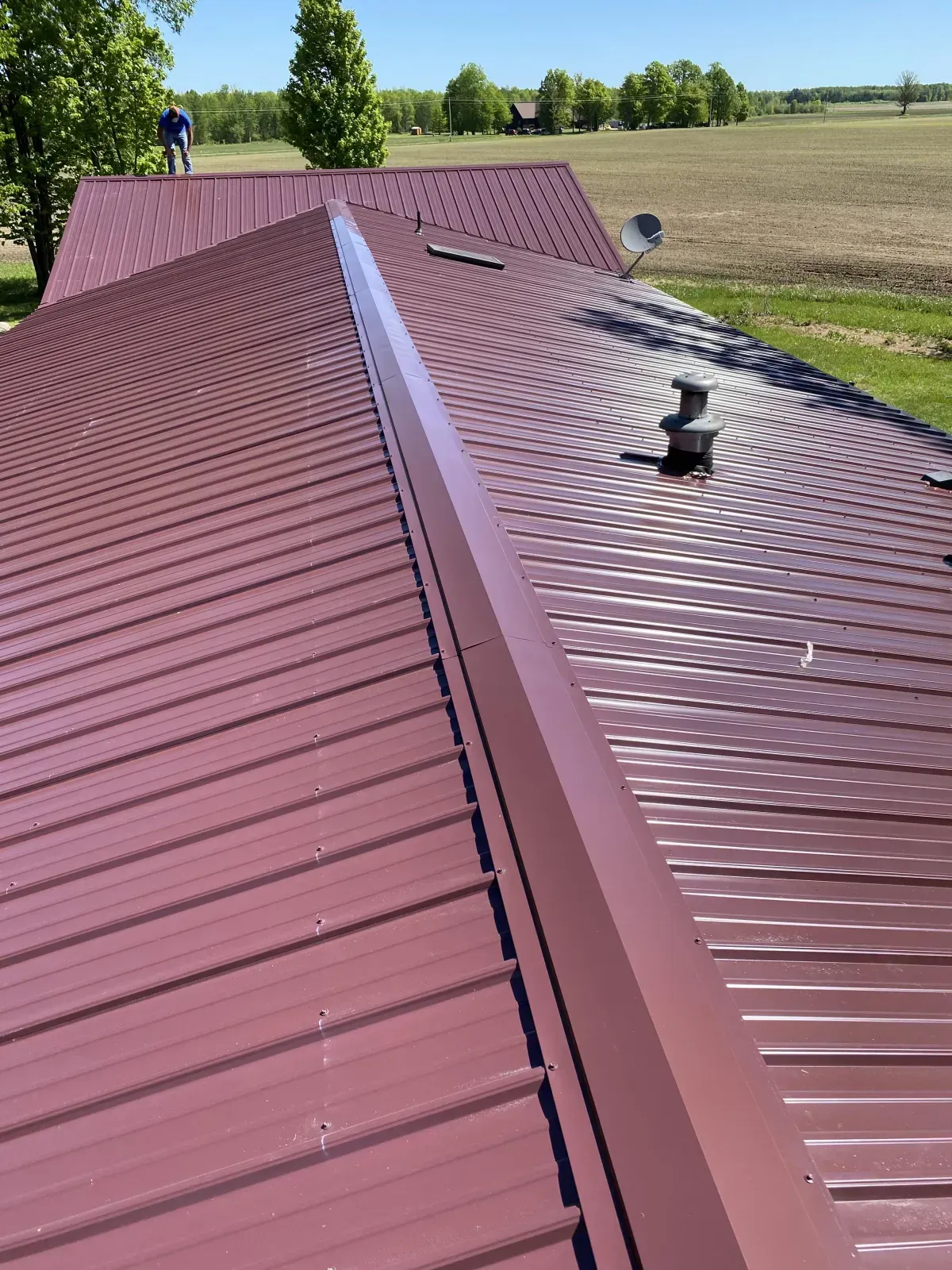 Red metal roof on a building with a central ridge, under a clear sky with trees and open field in the background