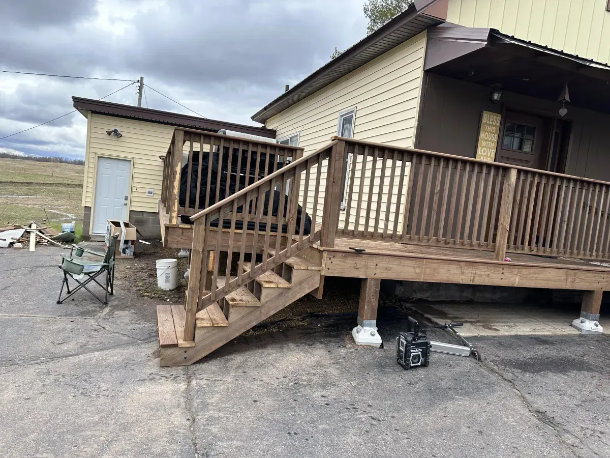 Weathered house porch with wooden stairs and railing on a gravel lot under cloudy skies