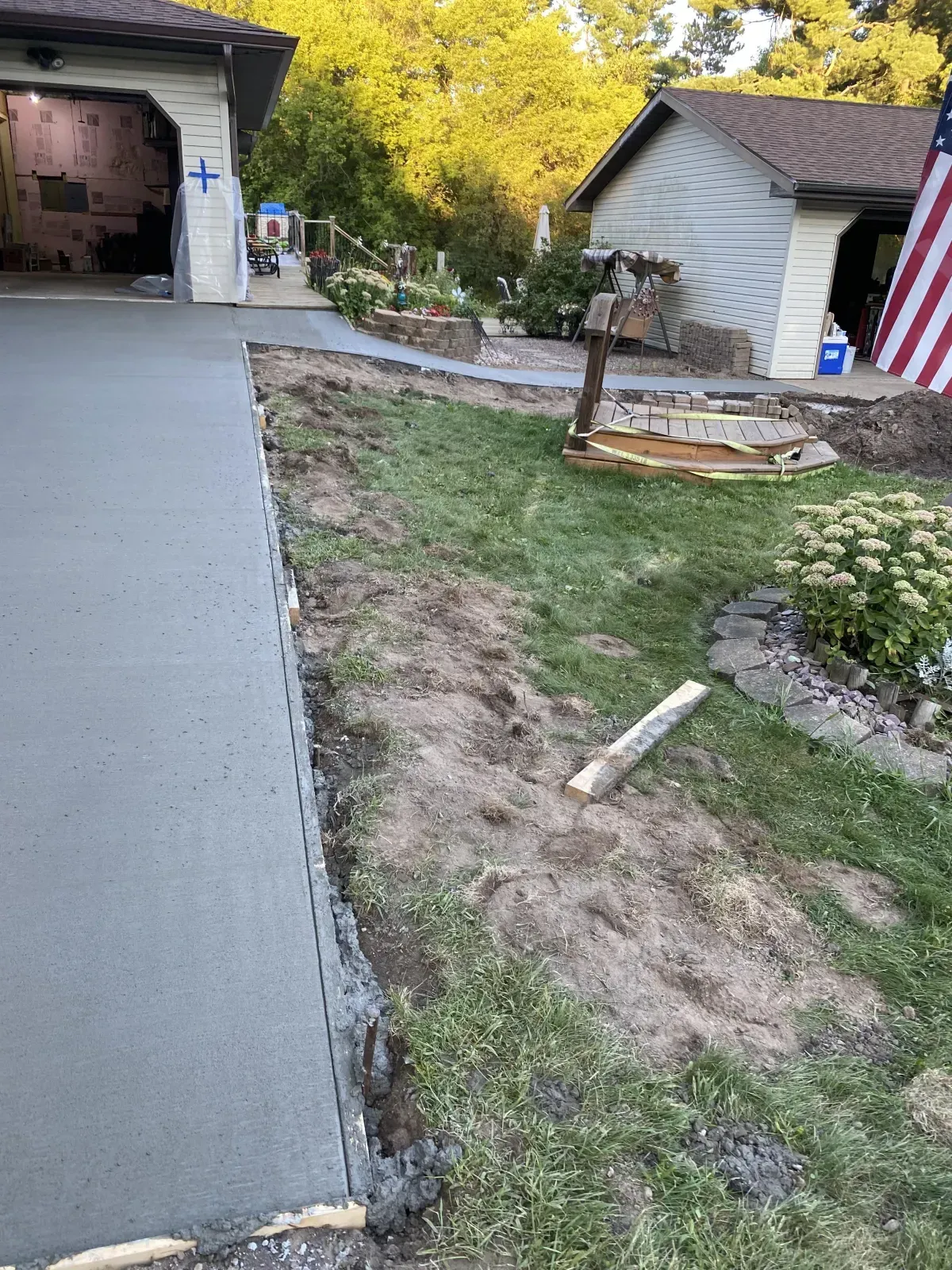 Concrete driveway beside a house, with a muddy grass strip and a small landscaped area near a garage.