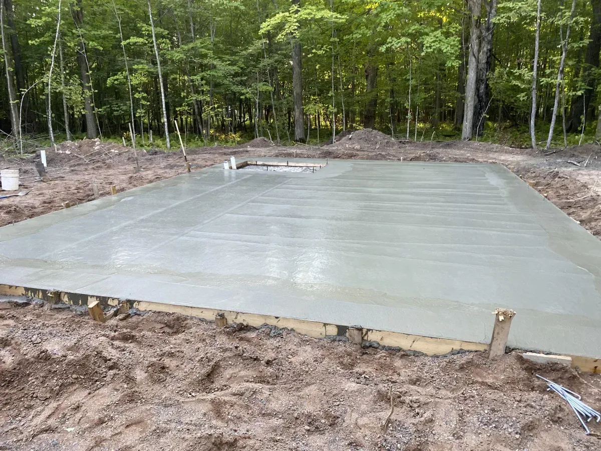 Fresh concrete slab foundation poured in a wooded lot, with forms and dirt around it