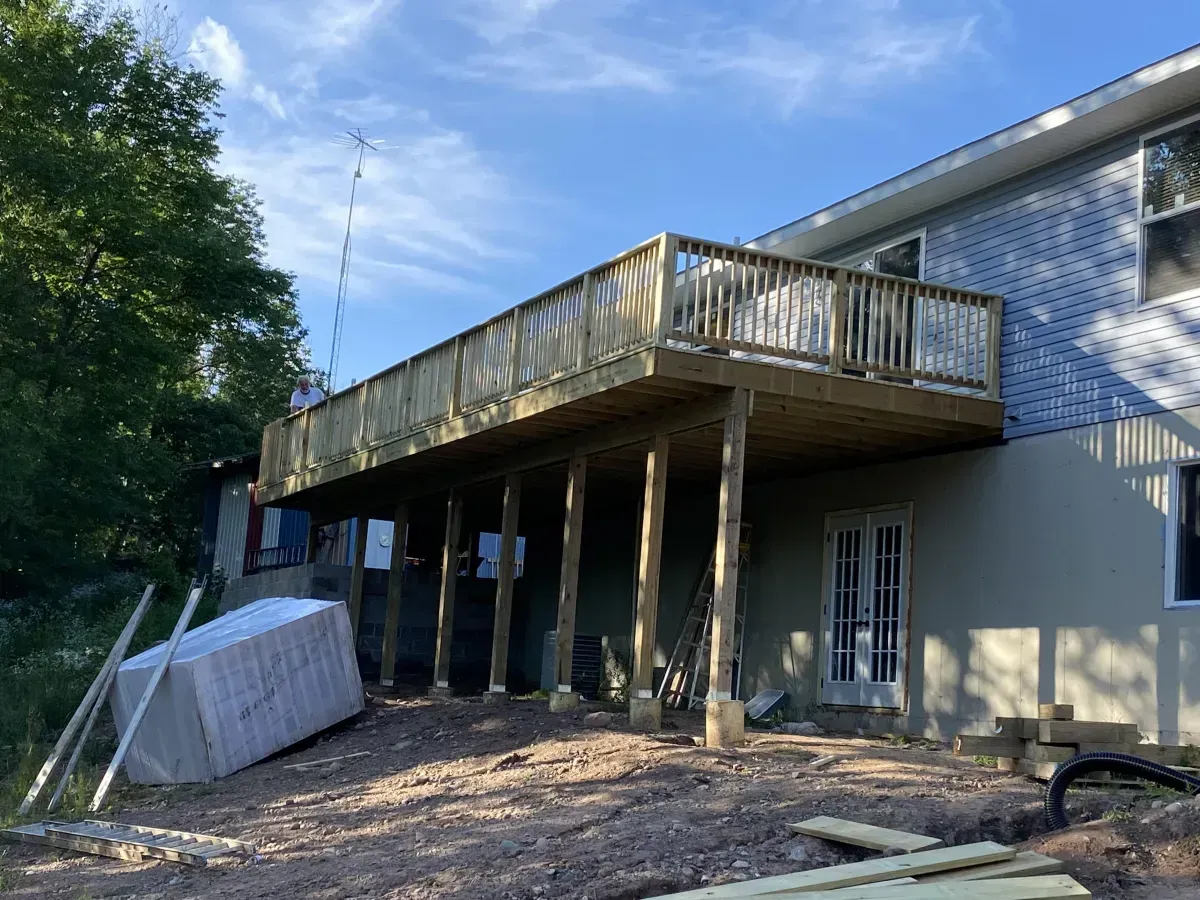 Raised wooden deck beside a blue house, with posts, railing, and debris below under a clear sky