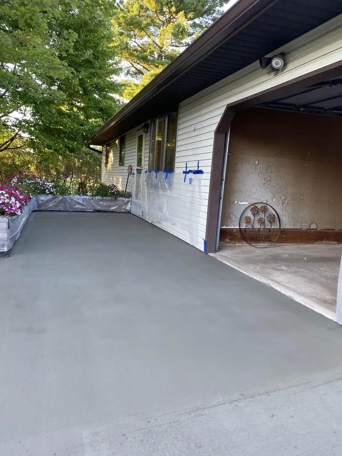 Freshly paved gray driveway beside a white house and open garage, with trees and flower planters along the side.