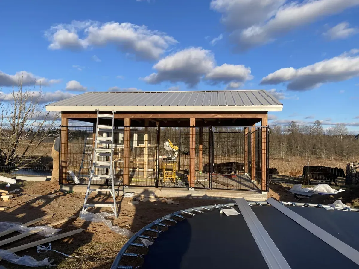 Small wooden shed under construction beside a driveway, with ladders, fencing, and snow patches.