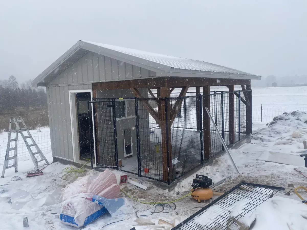 Snowy outdoor construction site with a small shed, metal framing, ladder, and tools scattered on the ground.