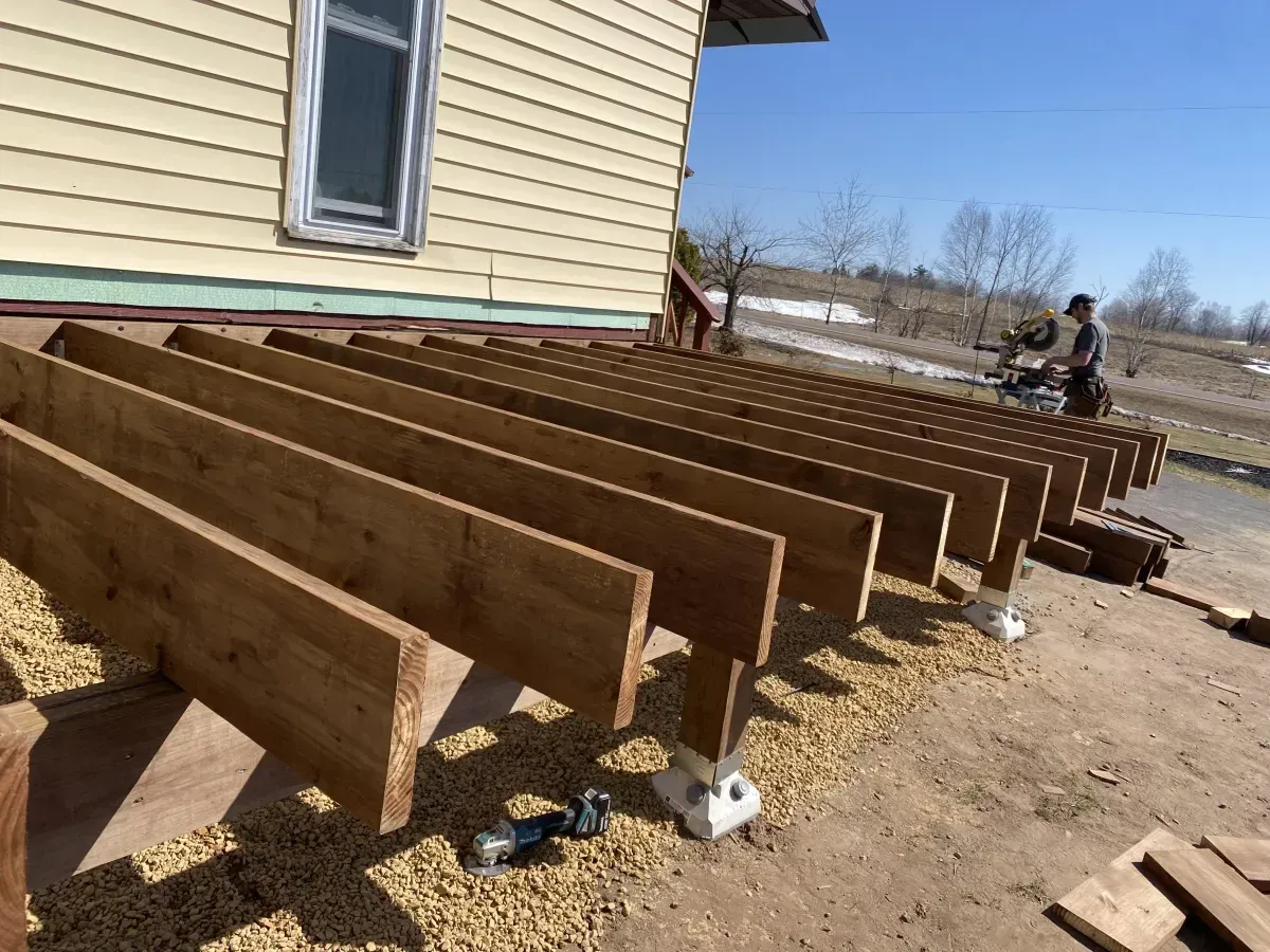 Wooden deck framing beams extending beside a beige house on a dirt lot, with workers in the background
