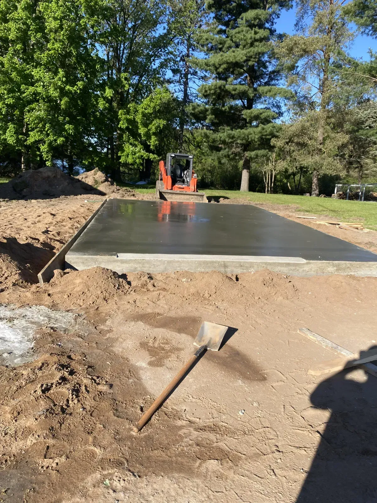 Fresh concrete slab being finished outdoors with a small excavator and shovel nearby.