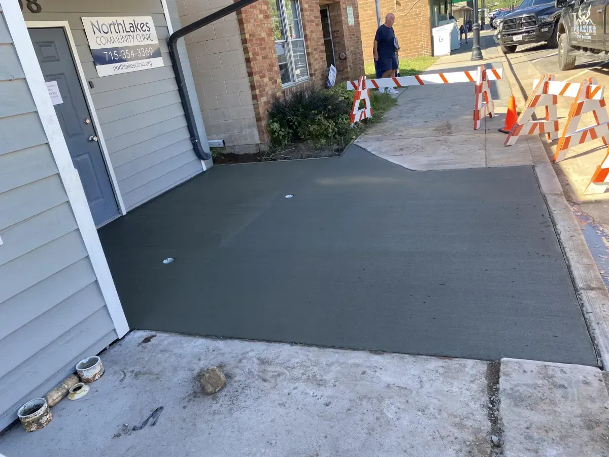 Fresh black concrete ramp beside a building, with orange barricades blocking the sidewalk ahead.