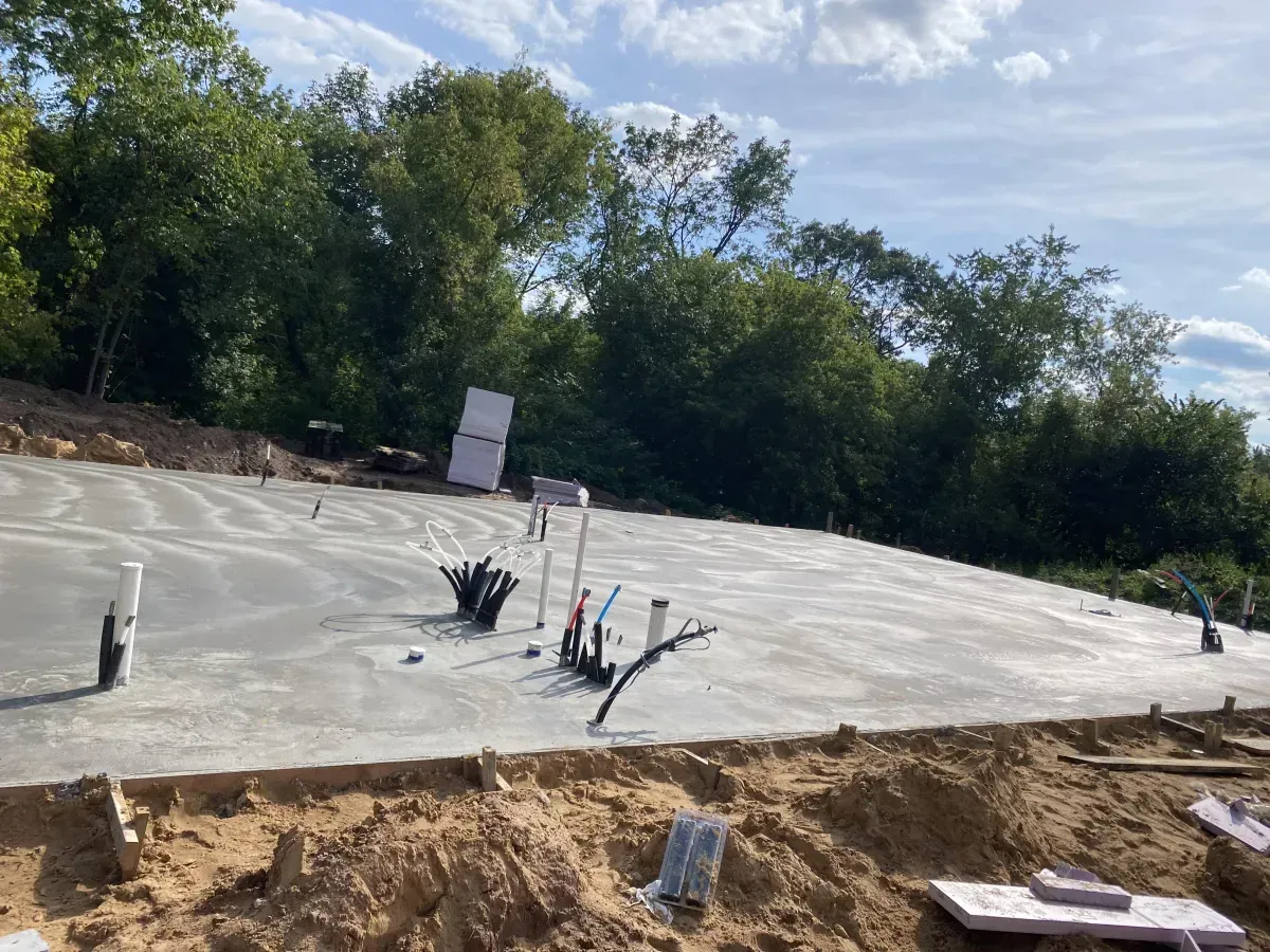 Construction site with concrete foundation slab, exposed plumbing pipes, and trees in the background