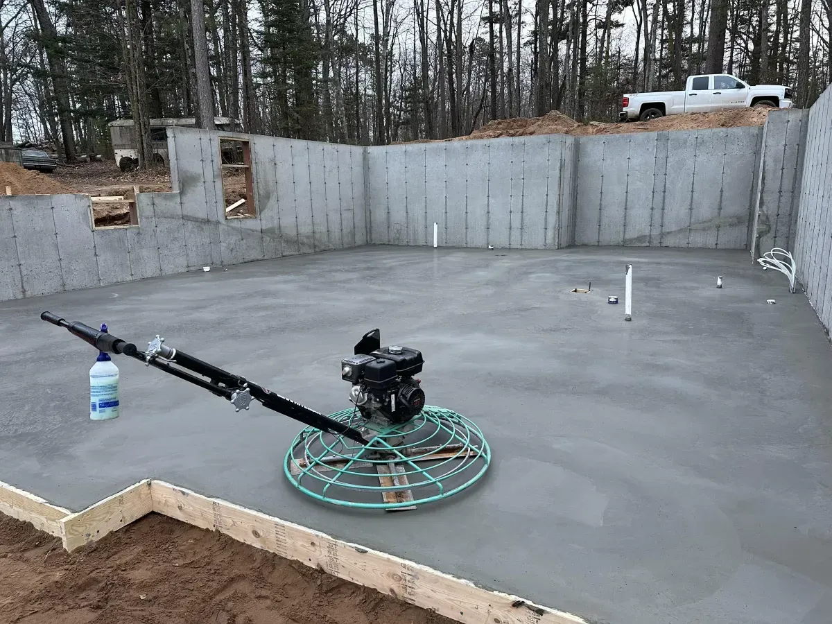 Concrete foundation slab with gray walls, spray rig, and a white truck in a wooded construction site
