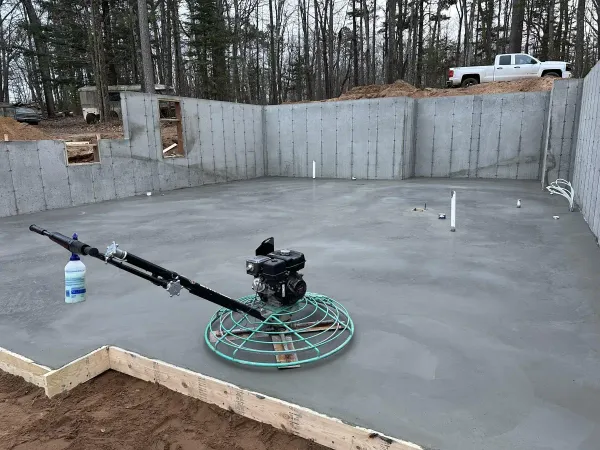 Concrete foundation slab with gray walls, spray rig, and a white truck in a wooded construction site